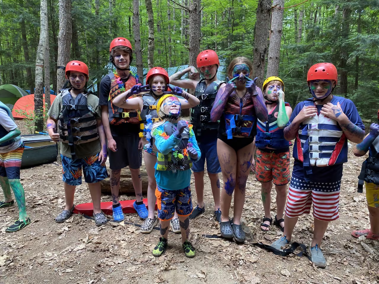 Junior campers with colorful face paint, helmets, and life jackets at their wooded campsite